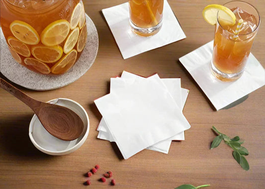Stack of white disposable beverage napkins on a wooden table with iced lemon tea glasses and a lemon-decorated pitcher