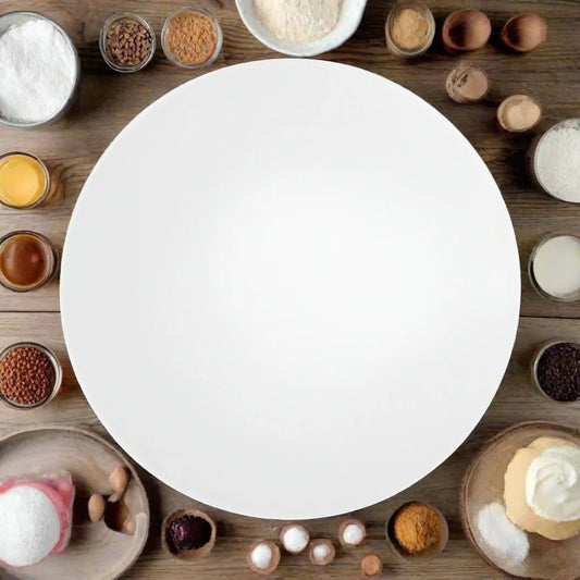 Empty white round cake board surrounded by various baking ingredients on a wooden surface