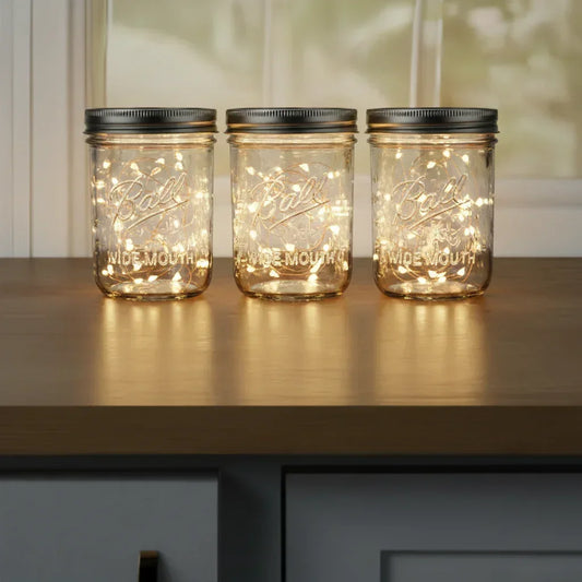 Three Ball wide mouth mason jars with glowing fairy string lights inside on wooden surface