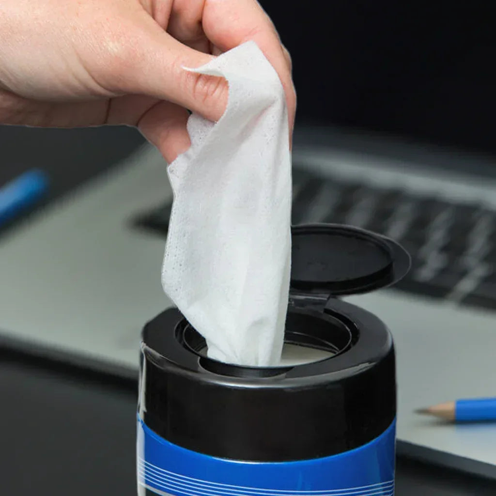 Hand pulling a white disposable cleaning wipe from a black and blue container on a desk with laptop and pencil