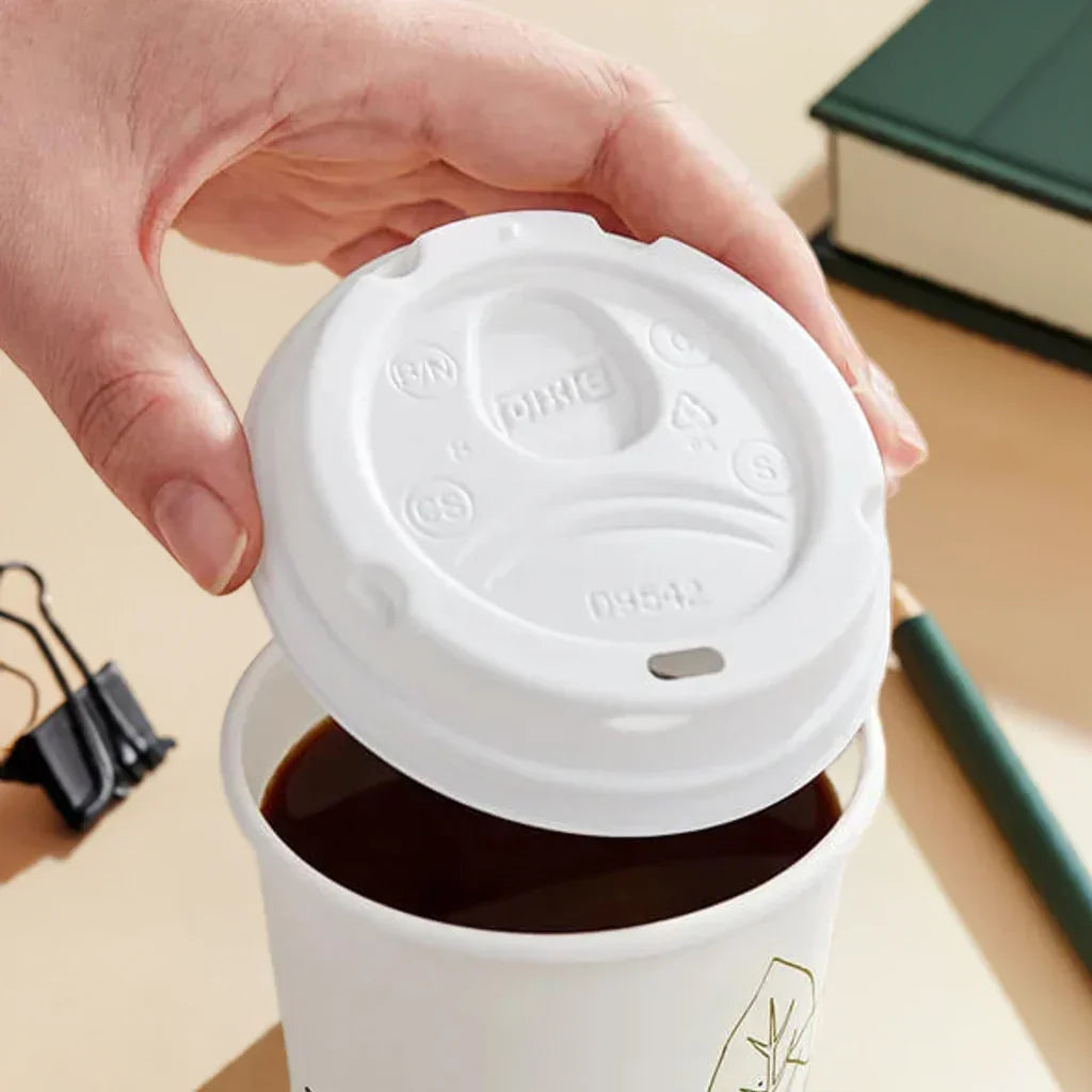 Hand lifting white disposable coffee cup lid to reveal black coffee inside, placed on desk with green pen, binder clip, and book
