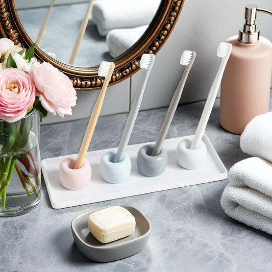 Four pastel-colored toothbrushes in matching holders on a tray with soap, lotion, white towels, and pink flowers on bathroom counter