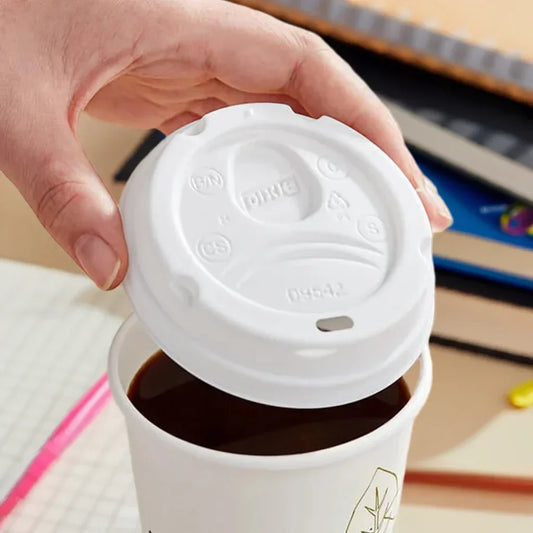Hand lifting white disposable coffee cup lid from cup filled with black coffee on office desk