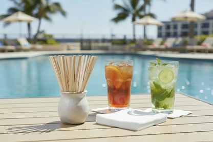 A bundle of Vanity Fair beverage napkins and bamboo stir sticks on a table with cocktails, with a swimming pool in the background.