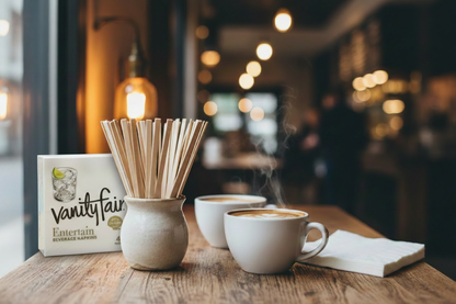 Coffee station setup in a modern cafe: a pack of premium Vanity Fair Entertain beverage napkins and eco-friendly bamboo cocktail stirrers in a vase are on a wooden table next to a latte.