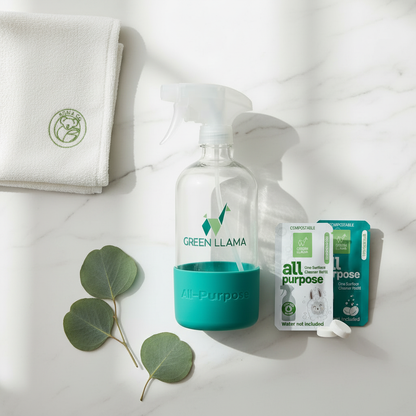 Eco-friendly cleaning kit with a Green Llama refillable glass spray bottle, cleaning tablets, and a white Koala Eco bamboo microfiber cloth on a marble surface.