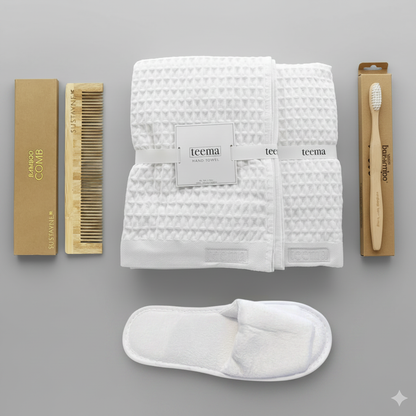 Curated luxury waffle bath towel spa gift bundle on white background, featuring a folded white organic Turkish cotton towel stack, plush velour Garnier-Thiebaut slippers, and eco-friendly bamboo essentials including a detangling comb and toothbrush.