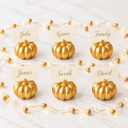 Six gold pumpkin place card holders with warm LED gold berry string lights on a pale marble background