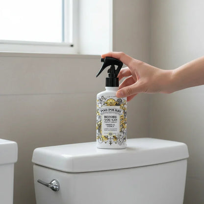 Hand holding a Poo-Pourri Before You Go toilet spray on a white toilet tank in a bright bathroom