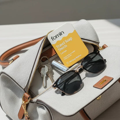 Beige handbag with brown handles containing sunglasses, keys, travel soap sheets, and lipstick on a white surface