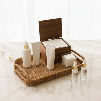 Natural woven vanity tray styled with wooden facial towel dispenser, clear square cotton pad holder, and minimalist skincare bottles on a marble countertop