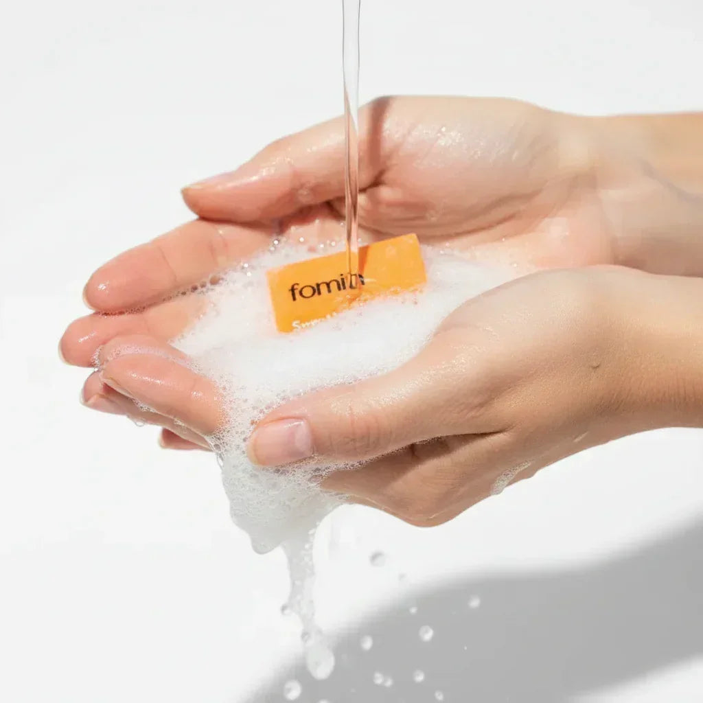 Hands holding foaming soap lather with water pouring over orange soap bar, bright white background