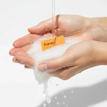 Hands holding foaming soap lather with water pouring over orange soap bar, bright white background
