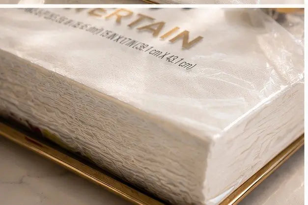 A close-up shot of a stack of premium white Vanity Fair Entertain paper napkins neatly placed on an elegant square glass tray with a polished gold metal rim on a white marble surface.