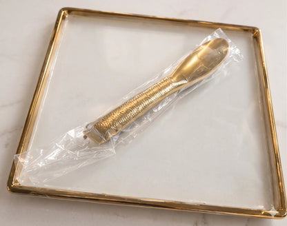 An empty minimalist square serving tray featuring a crystal-clear glass bottom and a modern shiny gold metallic border, displayed on a bright marble tabletop.